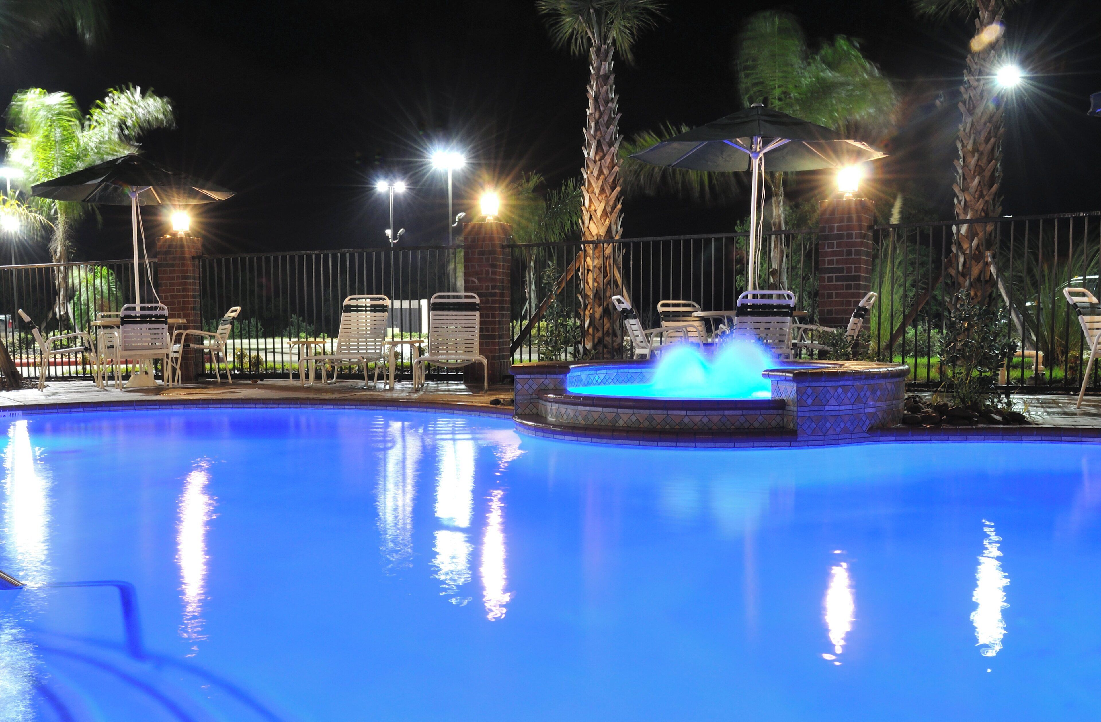 A swimming pool with a water feature, surrounded by lounge chairs and umbrellas, with a fence and palm trees in the background.