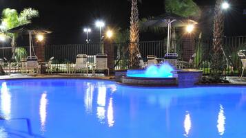 A swimming pool with a water feature, surrounded by lounge chairs and umbrellas, with a fence and palm trees in the background.
