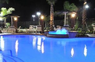 Piscina con fuente de agua, rodeada de sillas de descanso y sombrillas, con una cerca y palmeras al fondo.