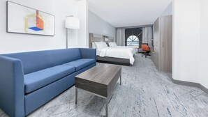 A modern hotel room with a blue sofa, a wooden coffee table, a bed with white linens, a desk, and a large window with curtains. - Holiday Inn Express Hotel & Suites Winnie by IHG (Winnie)