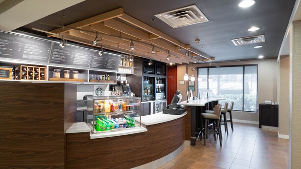 A modern bar area with a curved wooden counter, a menu board, and a seating area with a table and chairs. - Courtyard by Marriott Princeton (Princeton)