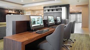 A modern office space with three computer workstations, each with a monitor, keyboard, and mouse. The room has a wooden desk, grey office chairs, and a carpeted floor. There are shelves with decorative items and a large window providing natural light.