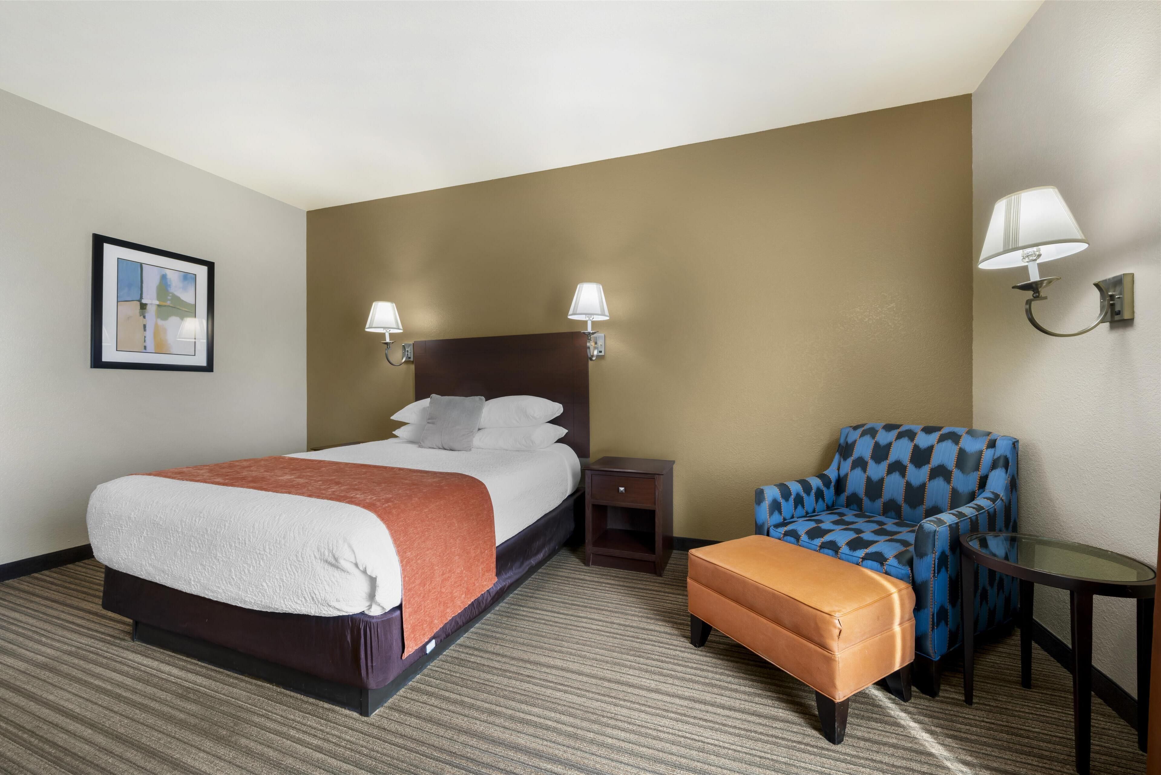 A hotel room with a bed, a chair, a small table, and two wall-mounted lamps.