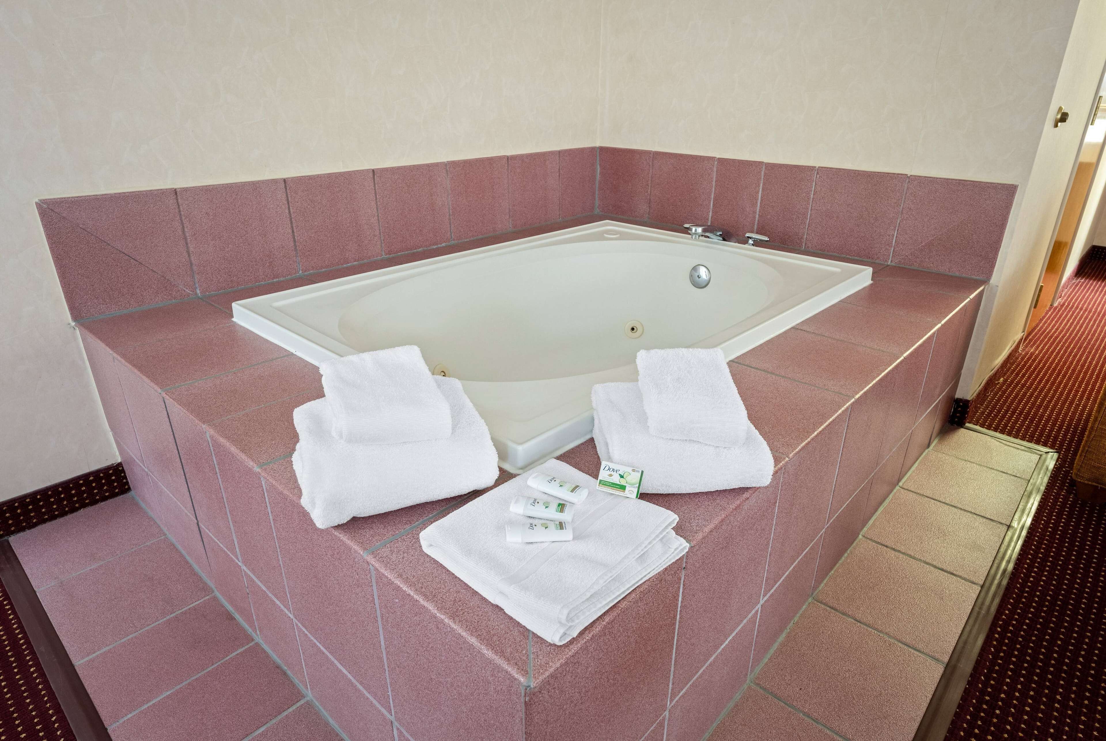 Eine große, rechteckige Badewanne mit Glastrennwand, umgeben von rosa gefliesten Wänden. Auf dem Boden liegen zwei weiße Handtücher und eine Flasche Dove Duschgel.