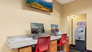 A hotel room with a desk, two red chairs, a desktop computer, a lamp, and an ATM.