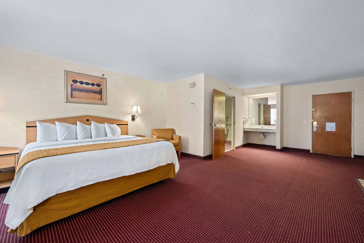 A hotel room with a large bed, a wooden headboard, a red carpet, a wall-mounted lamp, and a door leading to a bathroom.