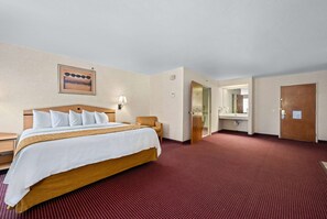 Una habitación de hotel con una cama grande, cabecera de madera, alfombra roja, una lámpara de pared y una puerta que da a un baño.