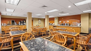 A hotel dining area with multiple tables and chairs, a kitchen area with appliances, and a clock on the wall.