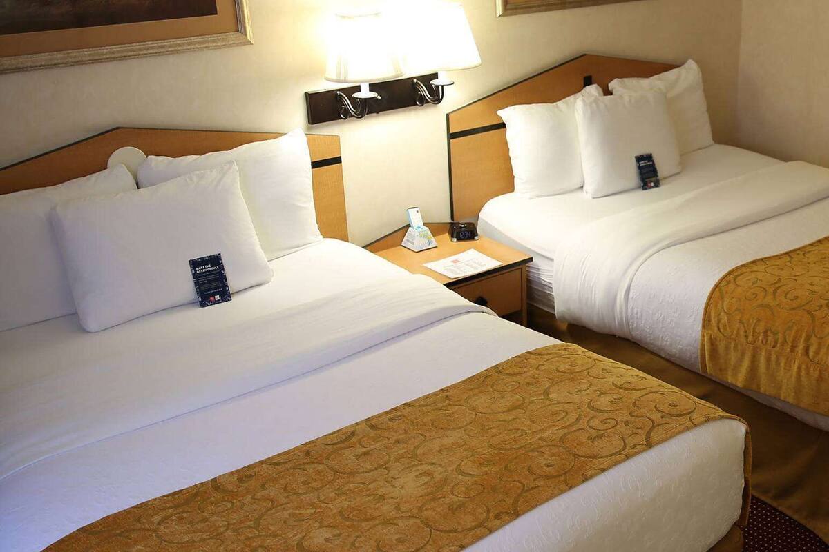 Two adjacent hotel beds with white linens, a bedside table with a lamp, and a wall-mounted light fixture.
