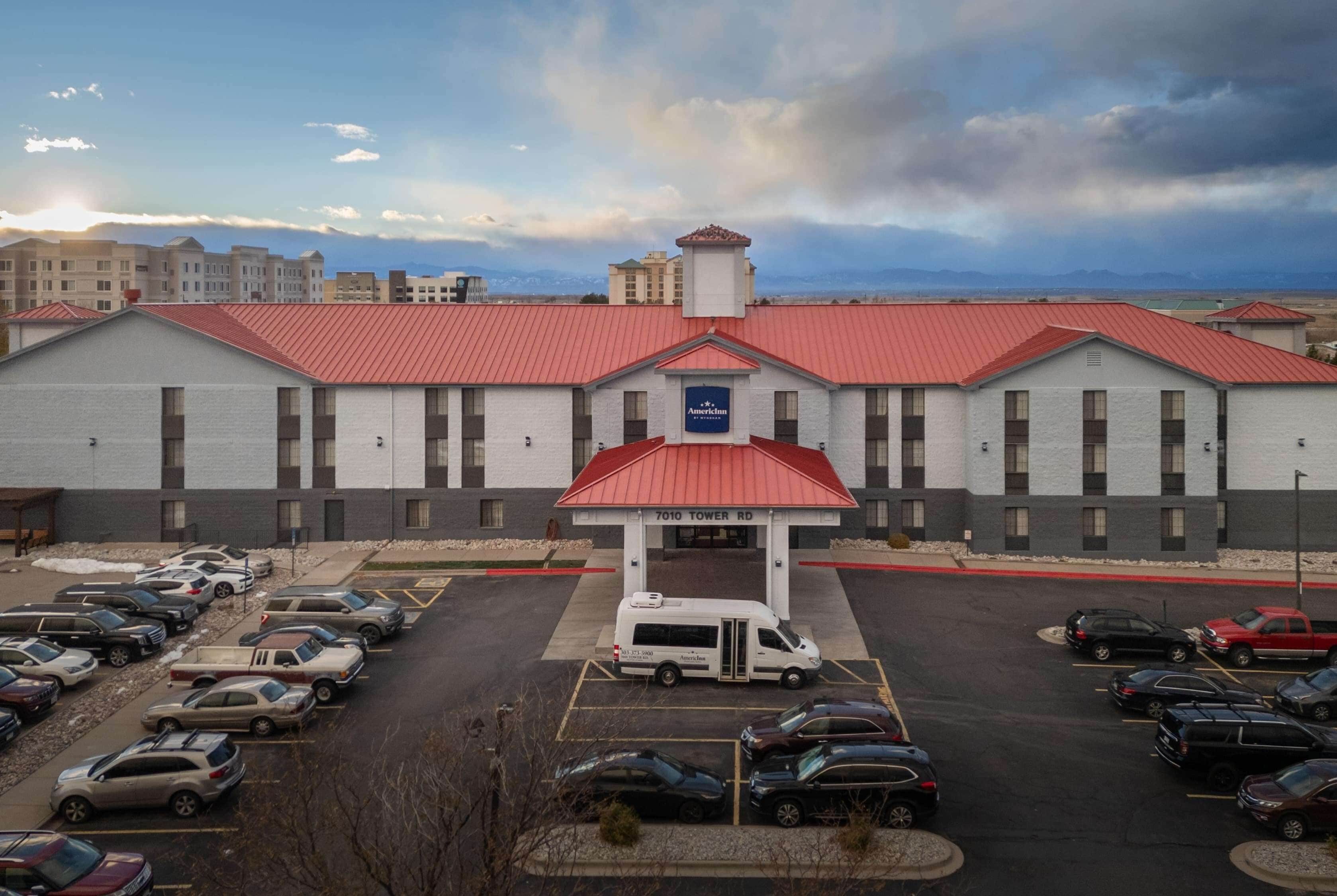 A hotel with a red roof and a sign reading "7010 Tower Rd." There is a parking lot with several cars and a white van in front.