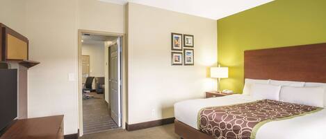 A hotel room with a bed, a desk, a chair, a lamp, and a door leading to another room.