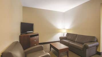 A hotel room with a brown leather sofa, a wooden coffee table, a TV on a wooden stand, and a lamp on a side table.