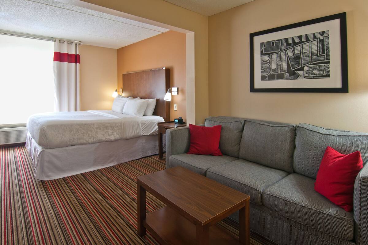 Une chambre d’hôtel avec un lit, un canapé, une table basse, un tableau encadré au mur et une fenêtre avec des rideaux.
