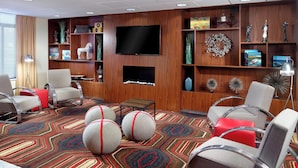 Eine Hotellobby mit modernem Design, inklusive eines großen Flachbildfernsehers, einem Kamin und verschiedener Dekorationselemente auf Regalen. Mehrere Stühle und ein gemustertes Teppich sind vorhanden.