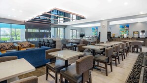 A modern restaurant with a mix of seating options including tables and sofas, a kitchen area, and large windows offering views of the outdoors. - Wingate by Wyndham Tinley Park (Tinley Park)