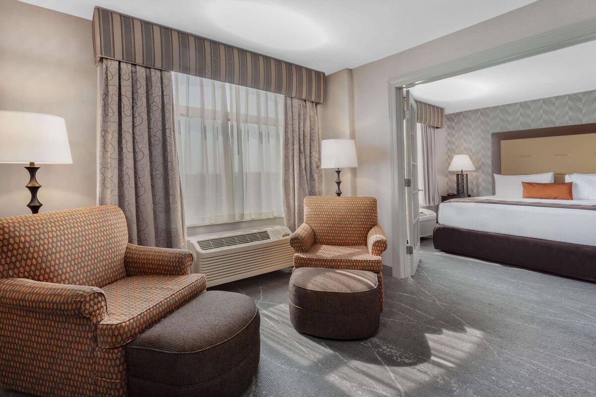 Ein Hotelzimmer mit einem großen Bett, zwei Sesseln, einem runden Hocker, einem Heizgerät und zwei Lampen.
