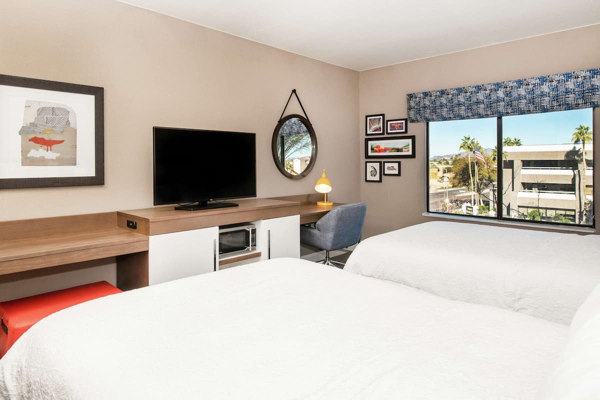 A hotel room with a bed, a desk with a TV, a small table with a lamp, and a window with a view of buildings and palm trees.