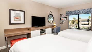Habitación de hotel con cama, escritorio con televisor, una mesita con lámpara y ventana con vista a edificios y palmeras.