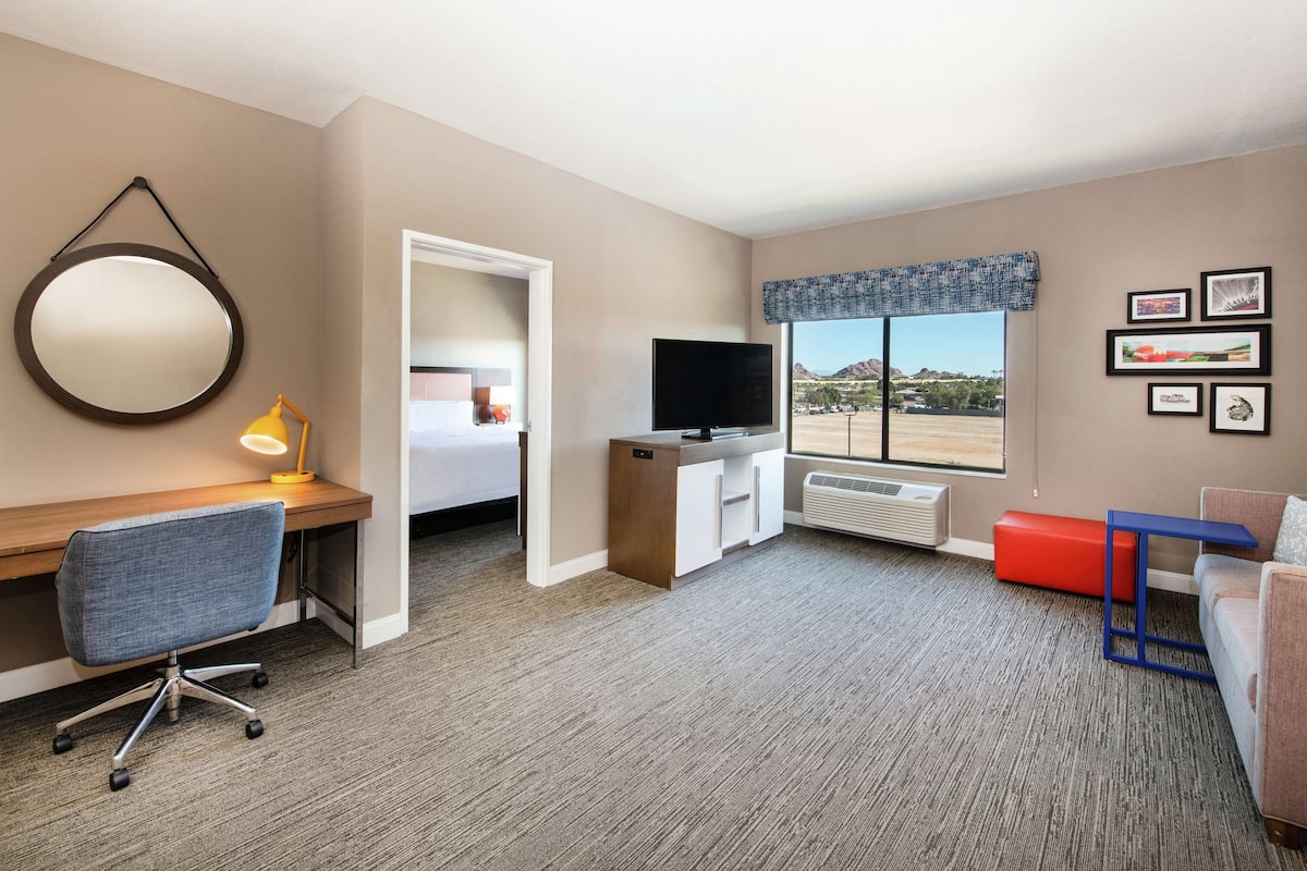 A hotel room with a desk, chair, TV, and a large window with a view.