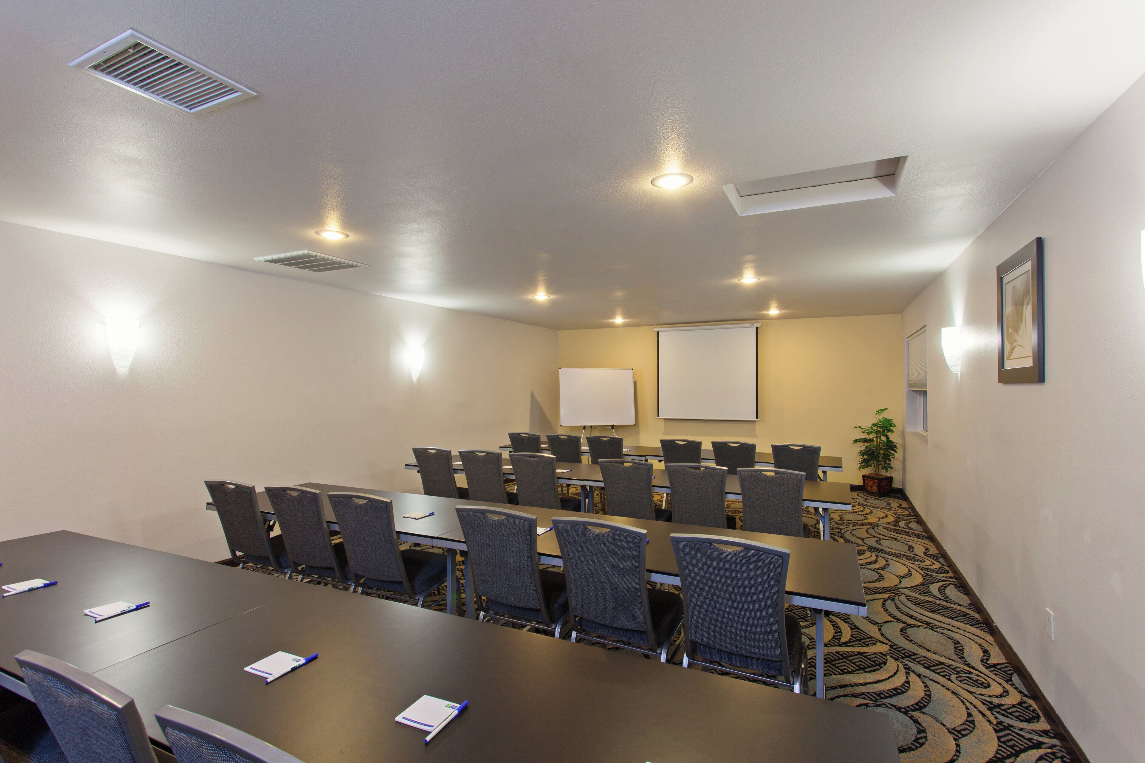 A conference room with a long table, multiple chairs, a whiteboard, and a projector screen.