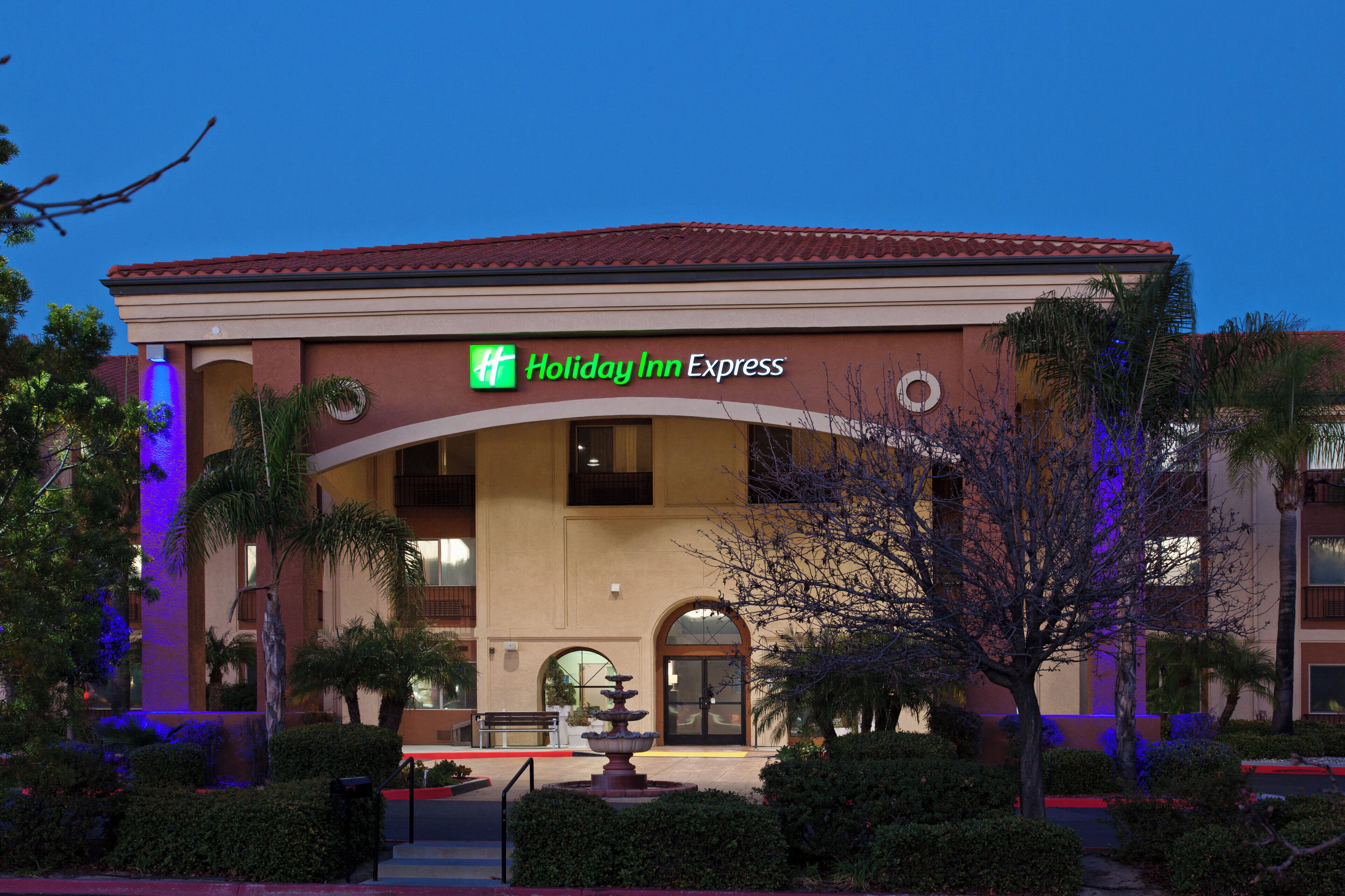 Un hotel Holiday Inn Express con techo de tejas rojas, una fuente en primer plano y palmeras a su alrededor.