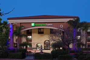Un hotel Holiday Inn Express con techo de tejas rojas, una fuente en primer plano y palmeras a su alrededor.