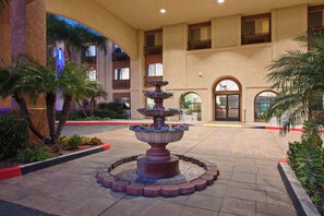 A courtyard with a central fountain, surrounded by palm trees and a building with multiple balconies.