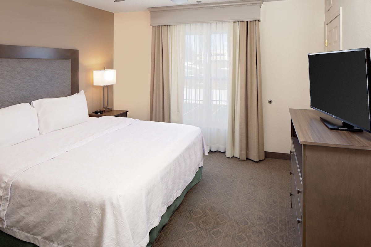 Habitación de hotel con una cama grande, una lámpara de noche, un televisor sobre un soporte de madera y una ventana con cortinas.