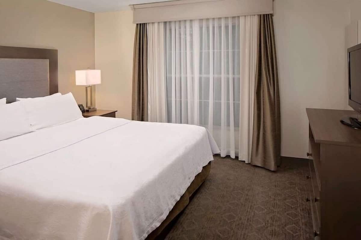 Habitación de hotel con una cama grande, una mesita de noche con lámpara, un televisor sobre un aparador de madera y una ventana con cortinas translúcidas.