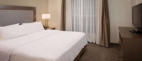 A hotel room with a large bed, a bedside table with a lamp, a TV on a wooden cabinet, and a window with sheer curtains.
