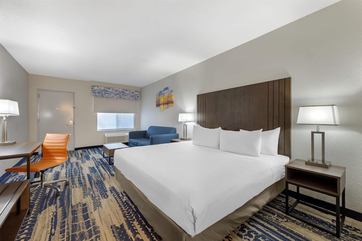 A hotel room with a large bed, a desk with a chair, a blue armchair, and a nightstand with a lamp.
