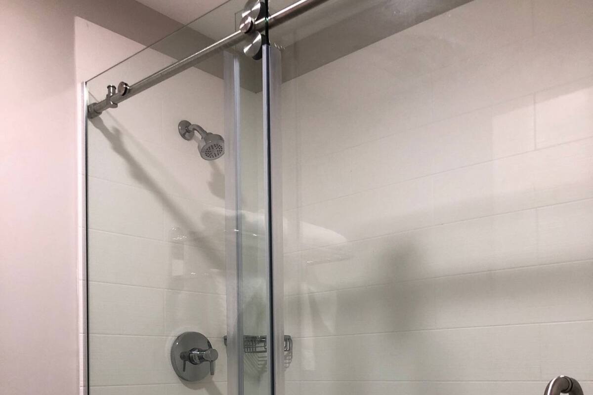 A glass-enclosed shower stall with a handheld showerhead, a wall-mounted faucet, and a toilet paper holder.