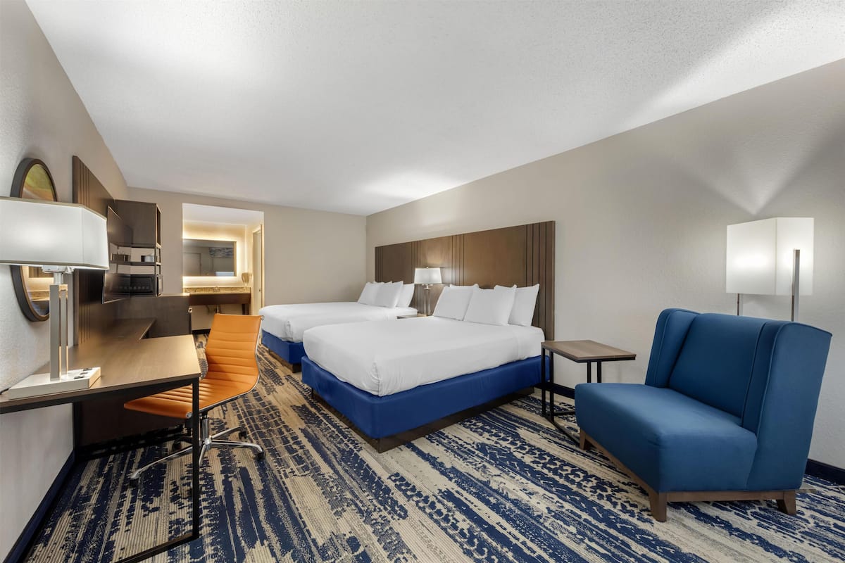 A hotel room with two beds, a desk, a chair, a small table, and a lamp.