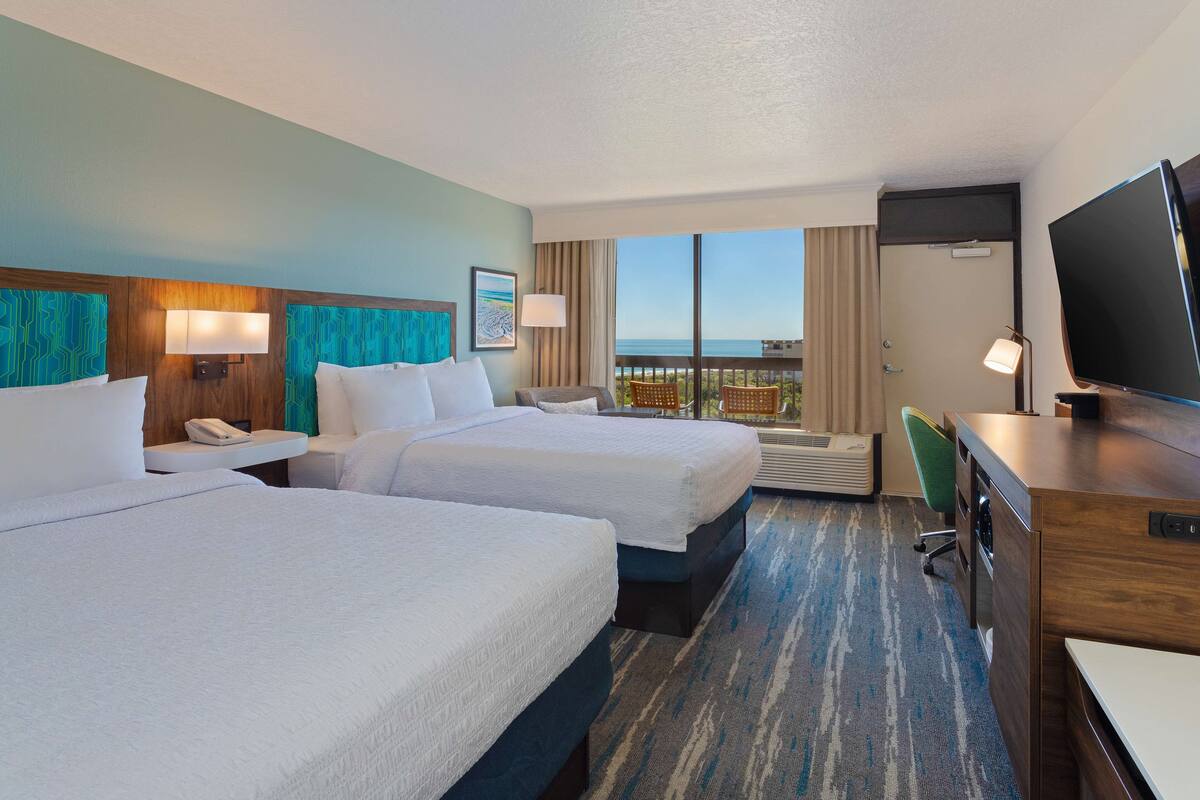 A hotel room with two beds, a desk, a TV, and a balcony with a view of the ocean.