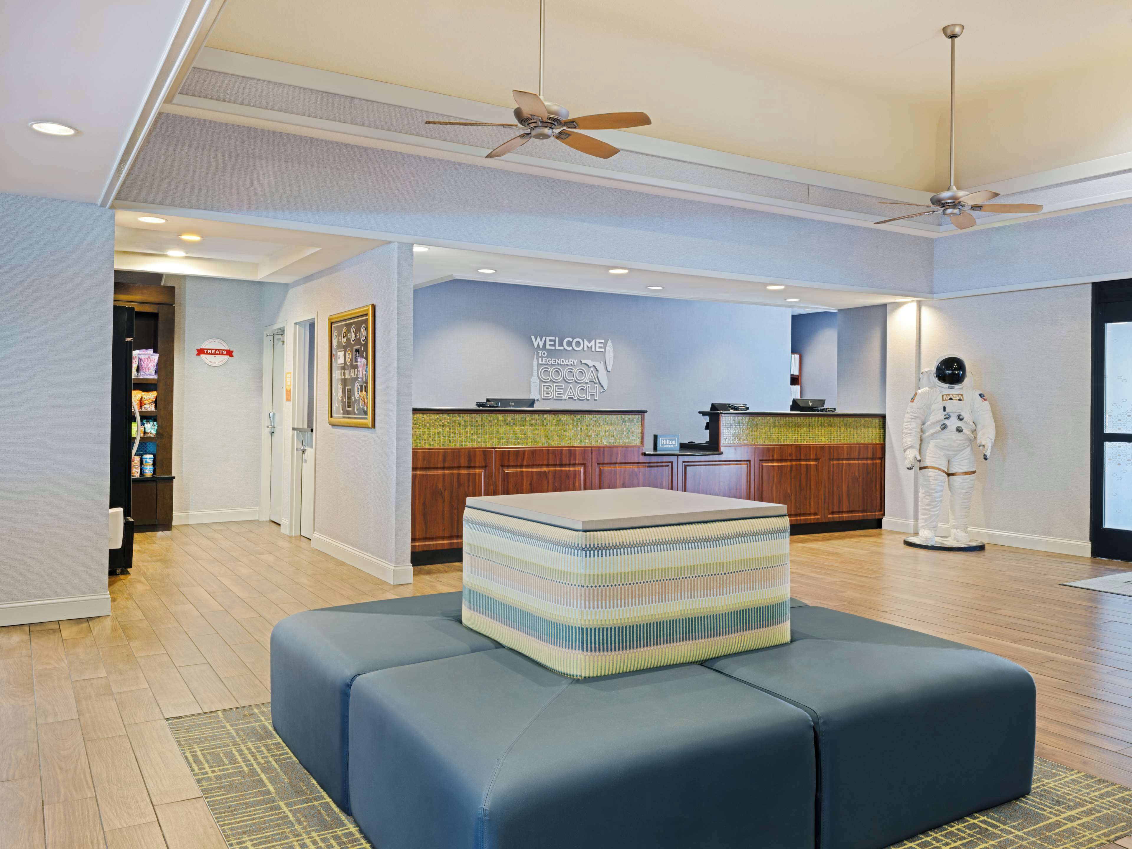 Un vestíbulo de hotel con una recepción, un cartel que dice "Welcome to Legendary Cocoa Beach" y una estatua de astronauta a tamaño real.