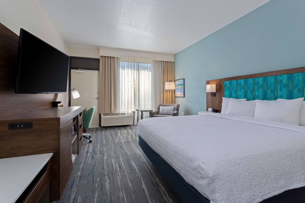 A hotel room with a large bed, a desk with a flat-screen TV, a chair, a desk lamp, and a window with curtains.