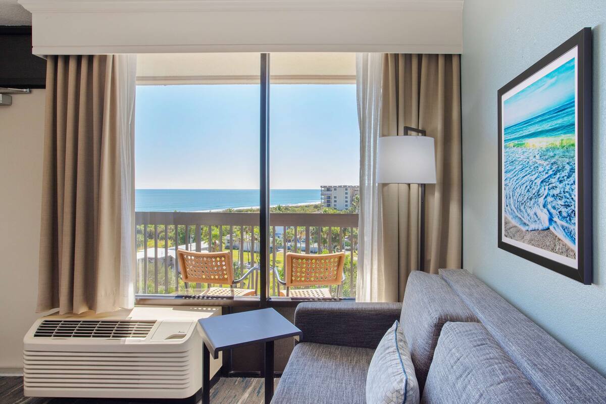 A hotel room with a balcony, a sofa, a table, and a wall-mounted air conditioner.