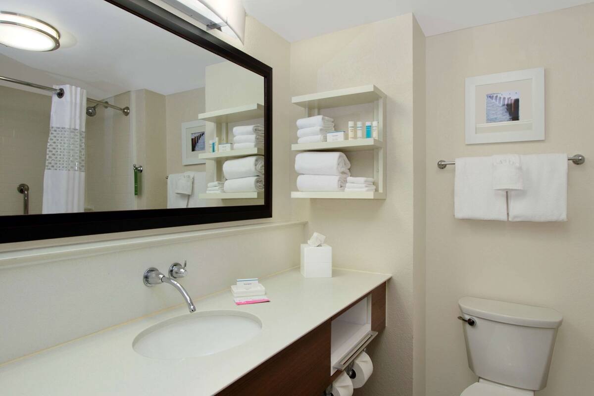 A hotel bathroom with a sink, mirror, towel rack, toilet, and shelves with toiletries.