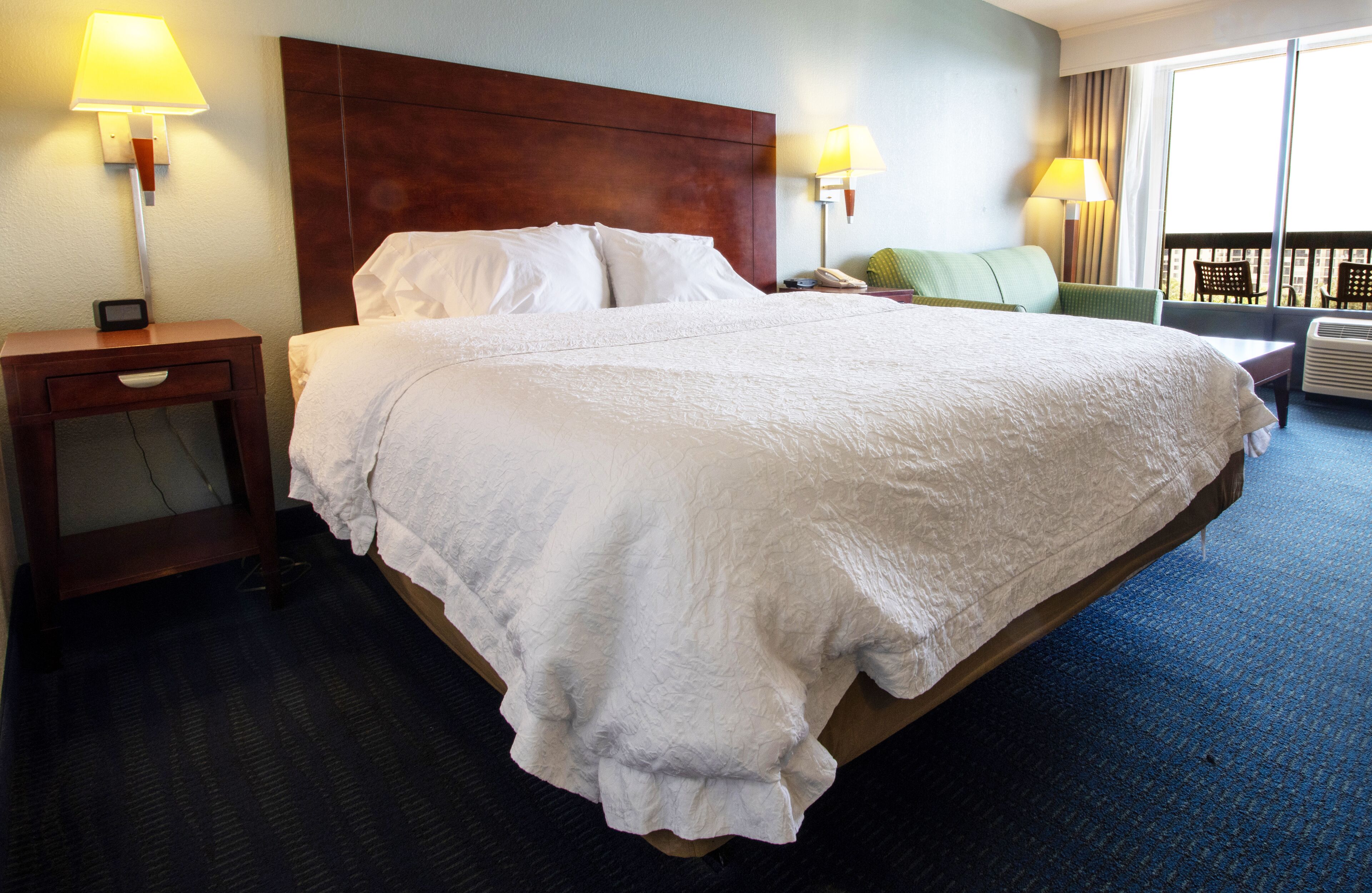 Een hotelkamer met een groot bed, twee nachtkastjes met lampen, een groene fauteuil en uitzicht op de stad door grote ramen.