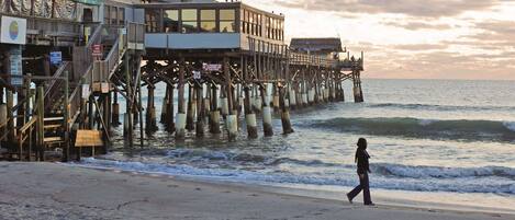 A pier extending into the water with a building on it, a person walking on the beach, and the ocean in the background.