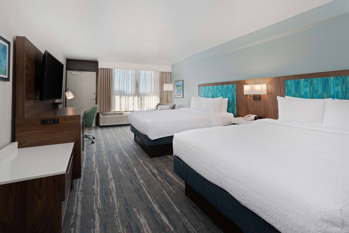 A hotel room with two beds, a desk, a TV, and a window with curtains.