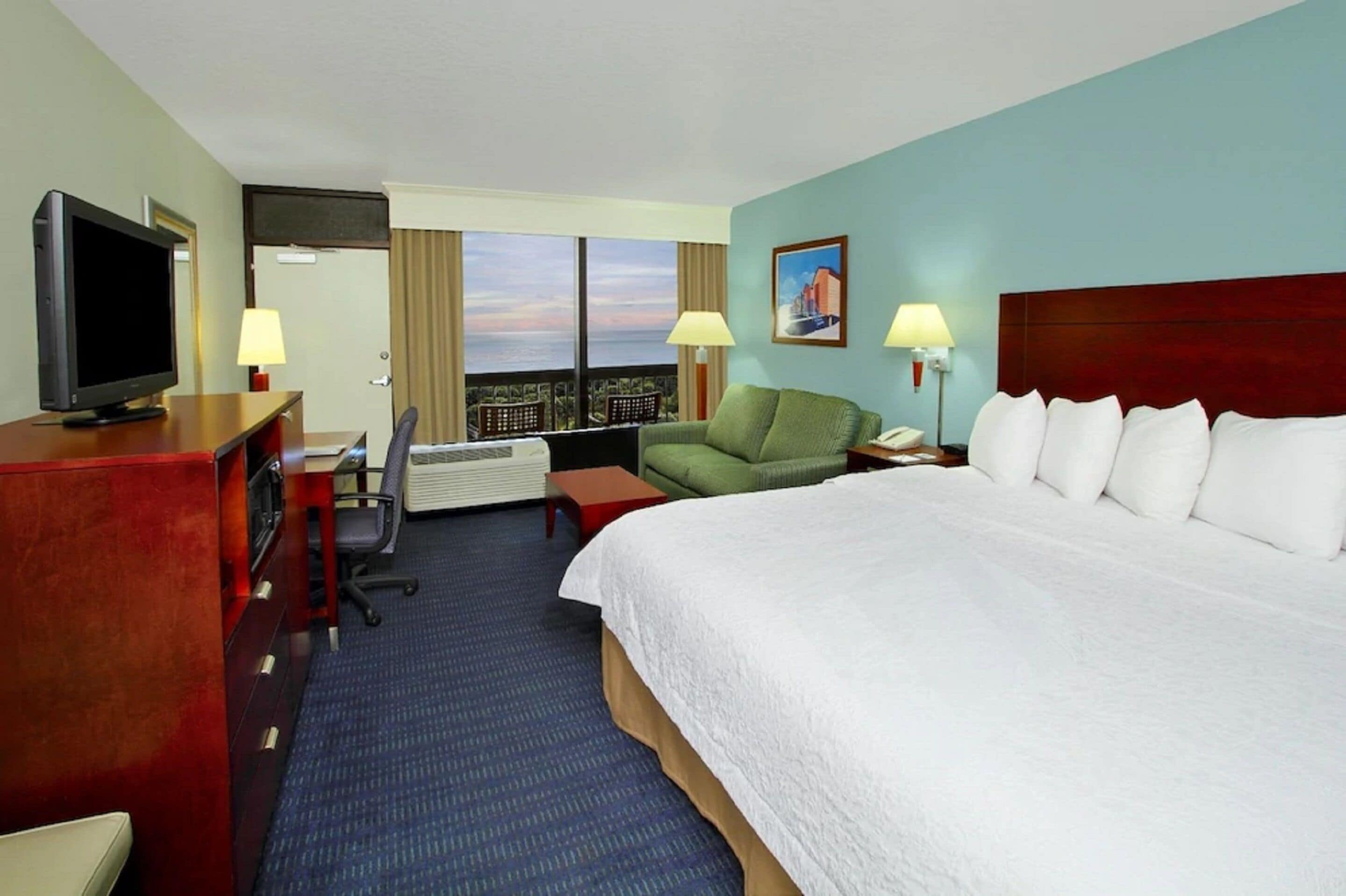 A hotel room with a large bed, a desk with a television, a chair, two lamps, and a view of the city through large windows.