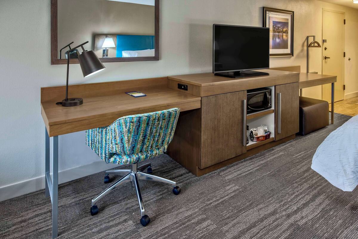 A hotel room with a wooden desk, a chair, a television, a microwave, and a small cabinet.
