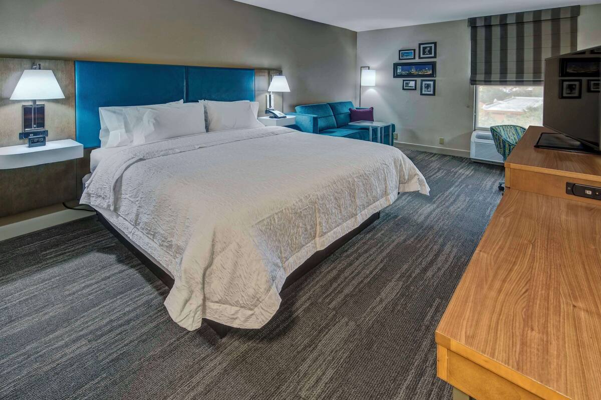 A hotel room with a large bed, bedside tables with lamps, a desk with a television, and a window with blinds.