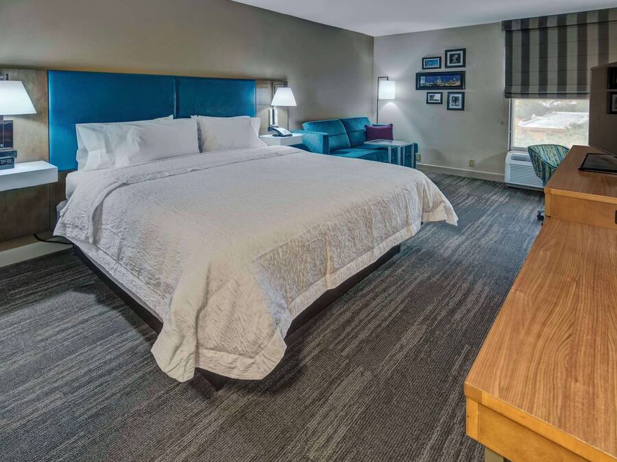 A hotel room with a large bed, bedside tables with lamps, a desk with a television, and a window with blinds.