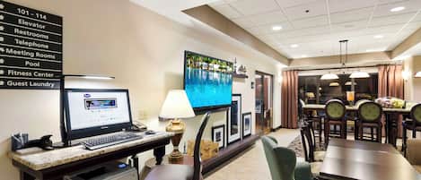 A hotel room with a desk, computer, printer, and a television. There is a sign with room numbers and amenities.