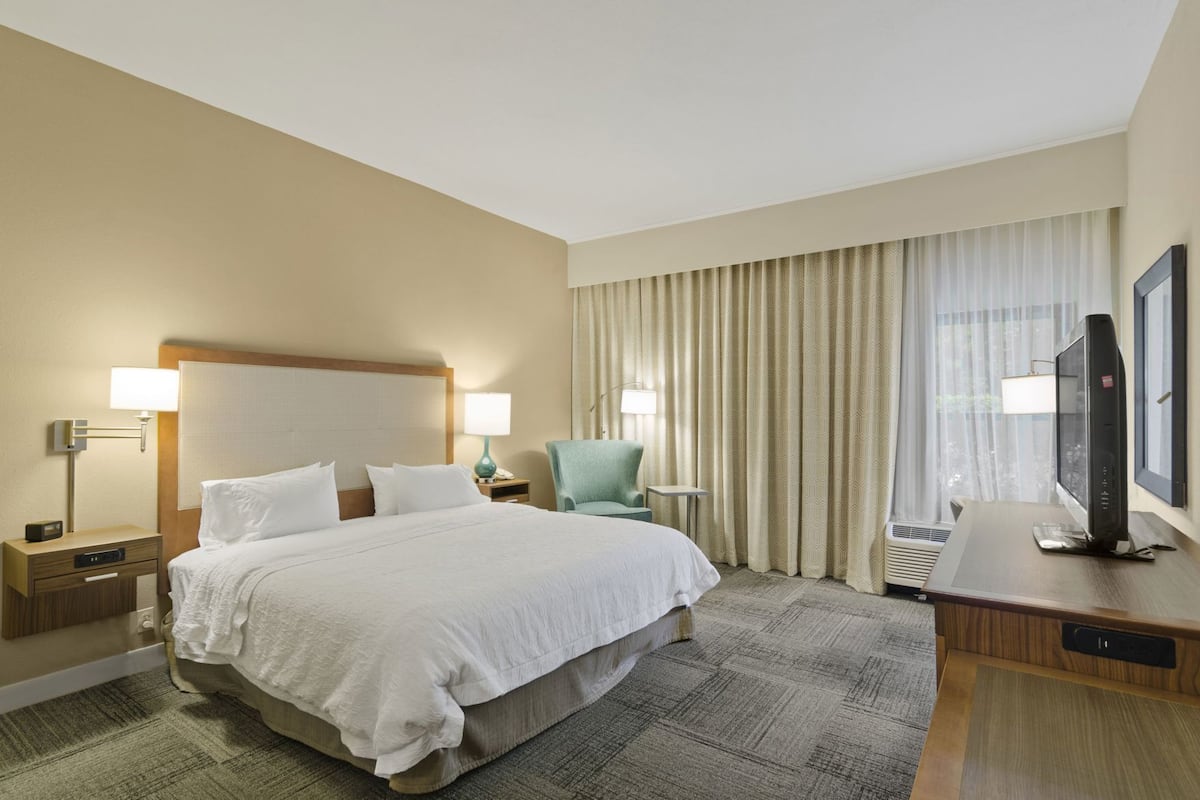 A hotel room with a large bed, two bedside tables with lamps, a chair, a desk with a television, and a window with curtains.