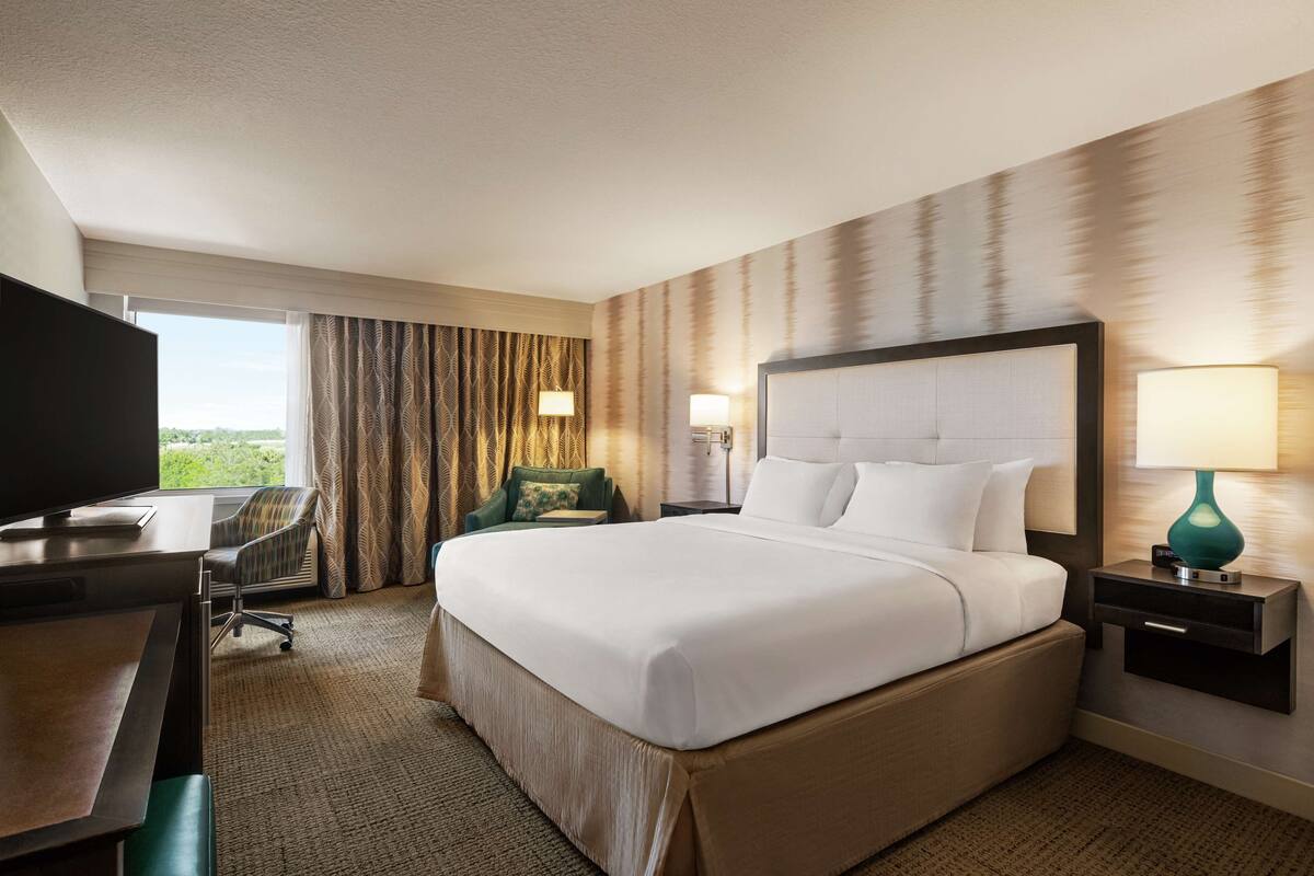 A hotel room with a large bed, a desk, a chair, a TV, and a window with curtains.