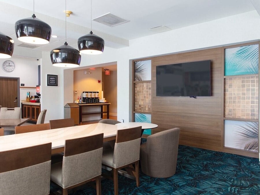 A modern hotel room with a large dining table, comfortable seating, a kitchen area, and a large TV screen.