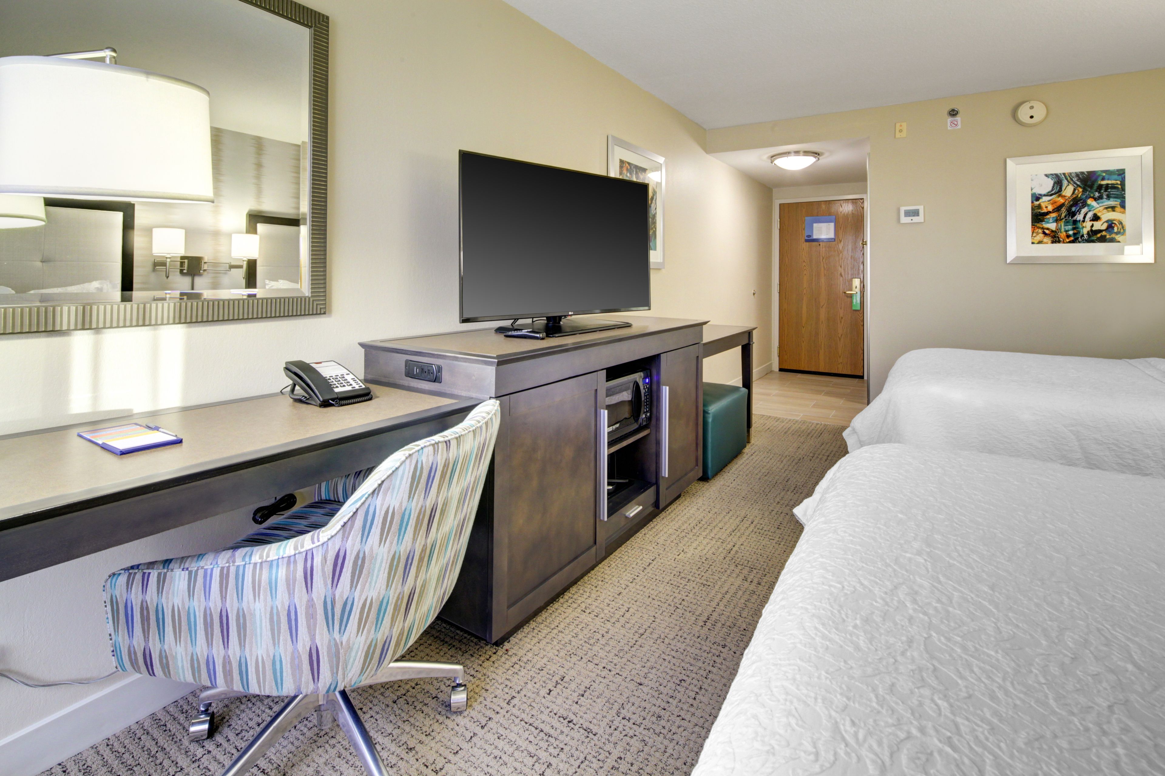 Quarto de hotel com cama, escrivaninha com computador, cadeira, televisão e espelho.